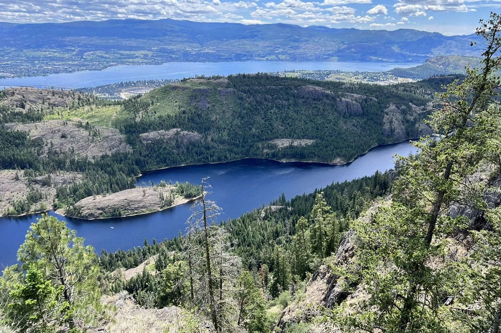 A breathtaking shot of the Rose Valley Reservoir with its deep blue hue, surrounded by rocky cliffs and forested slopes overlooking Okanagan Lake.